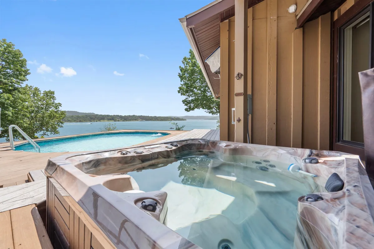 This exterior image beautifully showcases a shimmering swimming pool and an inviting hot tub, both set against a panoramic lake view. The deck is spacious, suggesting ample room for gatherings and relaxation, and the overall mood captures tranquility at sunset or dawn.