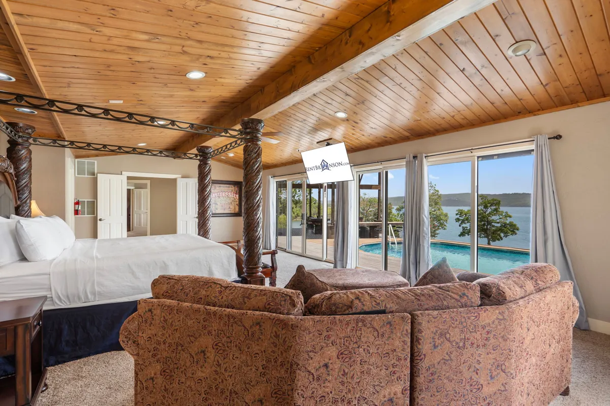 A spacious bedroom with a large bed, cozy sofa seating area, floor-to-ceiling windows overlooking the lake, and a warm wooden ceiling. The room exudes tranquility and luxury, showcasing elements of restful retreat with natural light pouring in and scenic lake views at the forefront.