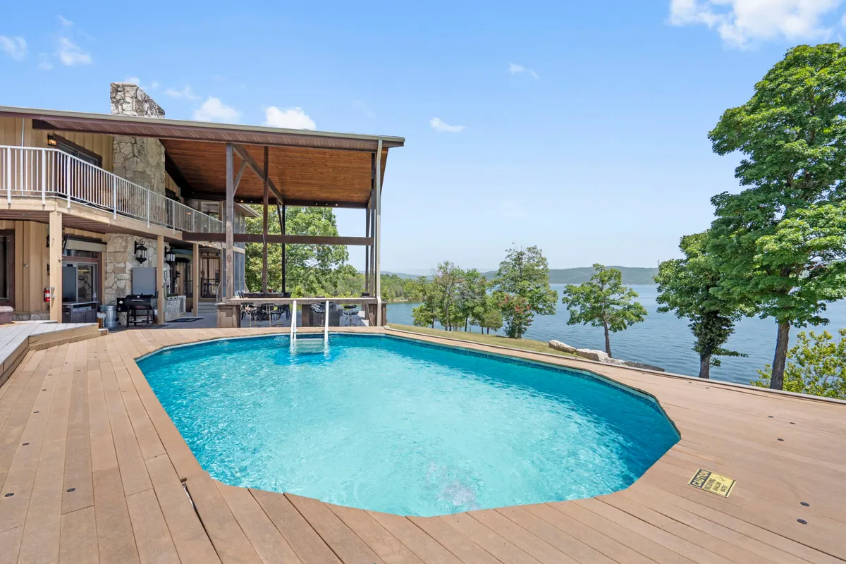 The image showcases a stunning exterior of a spacious lakefront home, with a sparkling private swimming pool in the foreground, a large deck area perfect for gatherings, and sweeping panoramic views of Table Rock Lake stretching into the distance. The scene is bathed in warm, inviting light, evoking relaxation and fun.