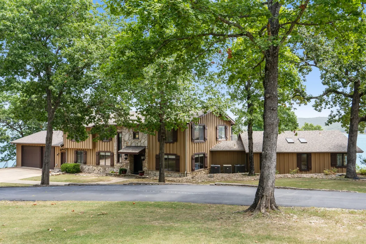 This exterior image showcases the house with a clear view of the large driveway, mature trees, and the lake in the background. The perspective highlights the expansive parking area suitable for multiple boats and trailers, as well as the property’s proximity to the water.
