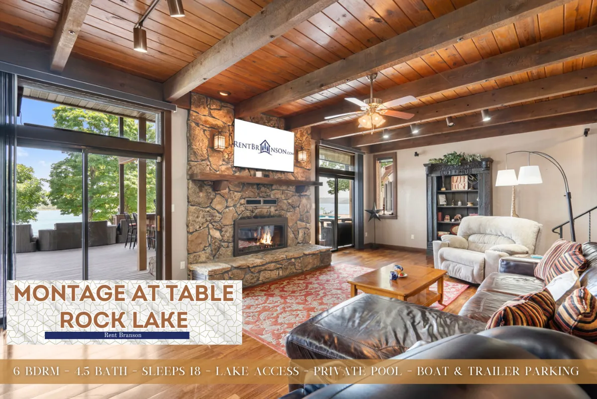 This image displays a warm, open-concept living room featuring a prominent stone fireplace, comfortable seating, a TV mounted above the fireplace, and large windows framing a spectacular view of Table Rock Lake. The space radiates rustic charm and provides a cozy setting ideal for family gatherings and relaxation after a day on the water.
