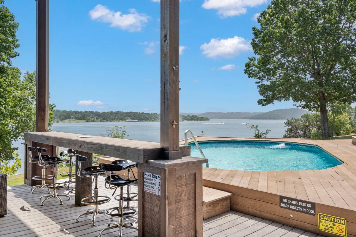 An outdoor scene with a swimming pool, bar area, and stunning lake views in the background. The setup encourages socializing and leisure, with areas for swimming, sunbathing, and enjoying drinks or casual meals by the water.