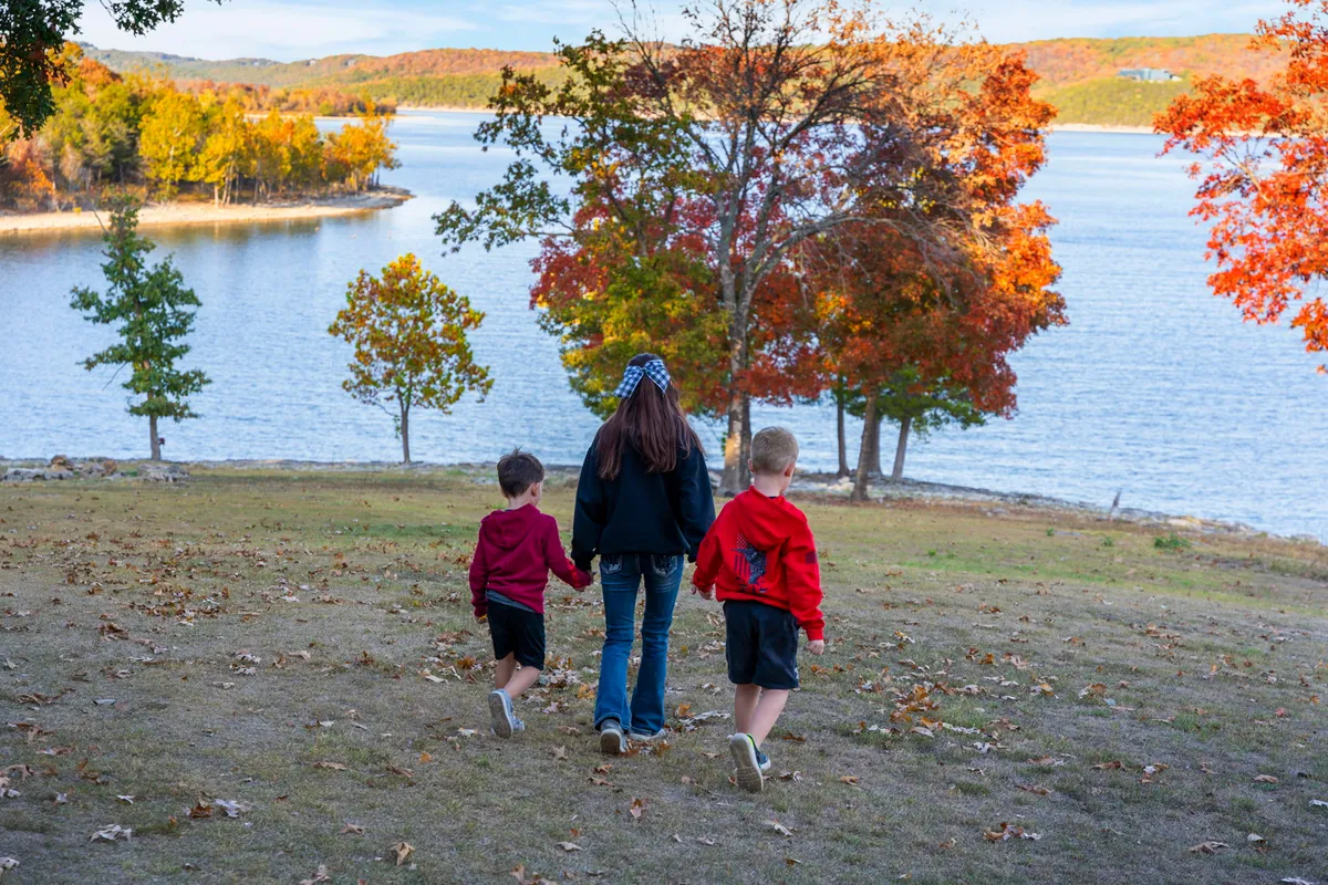 Children stroll along a lakeside path surrounded by colorful trees, with a water body glimmering in the background. The mood is playful, evoking the spirit of discovery as the children collect leaves and observe wildlife.