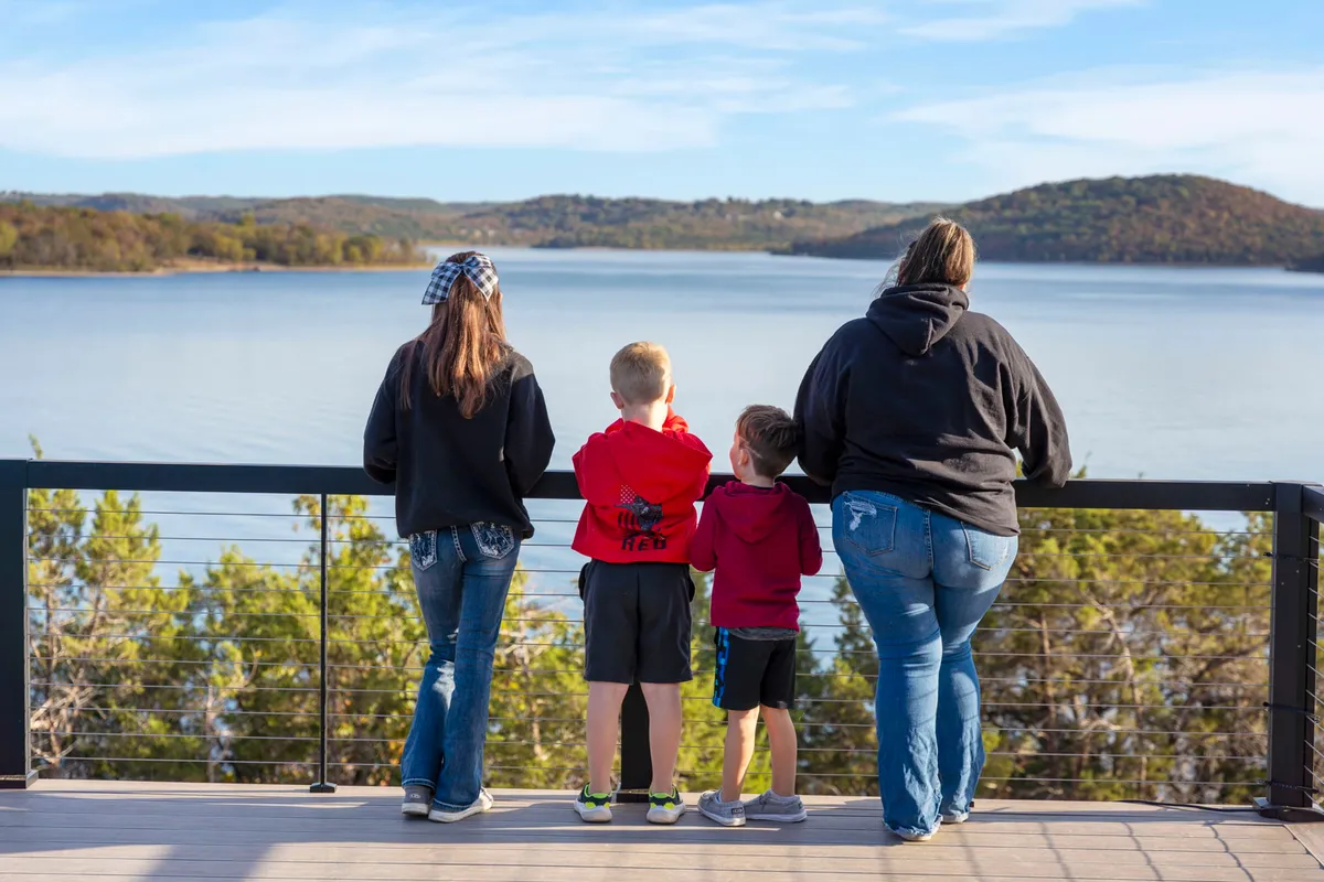 A family stands by the edge of a tranquil lake, looking out over calm water with distant, rolling hills. The scene captures children playing and adults relaxing, all set against the reflective surface of the lake and a serene, natural backdrop.